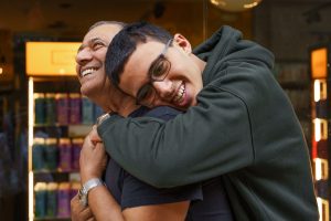 A teen boy leans over to hug his father, who to be smiling at him.