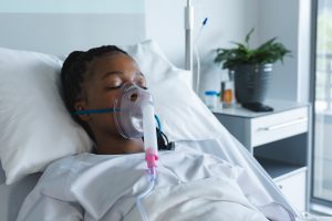 African american female patient with oxygen mask, lying on bed in hospital room